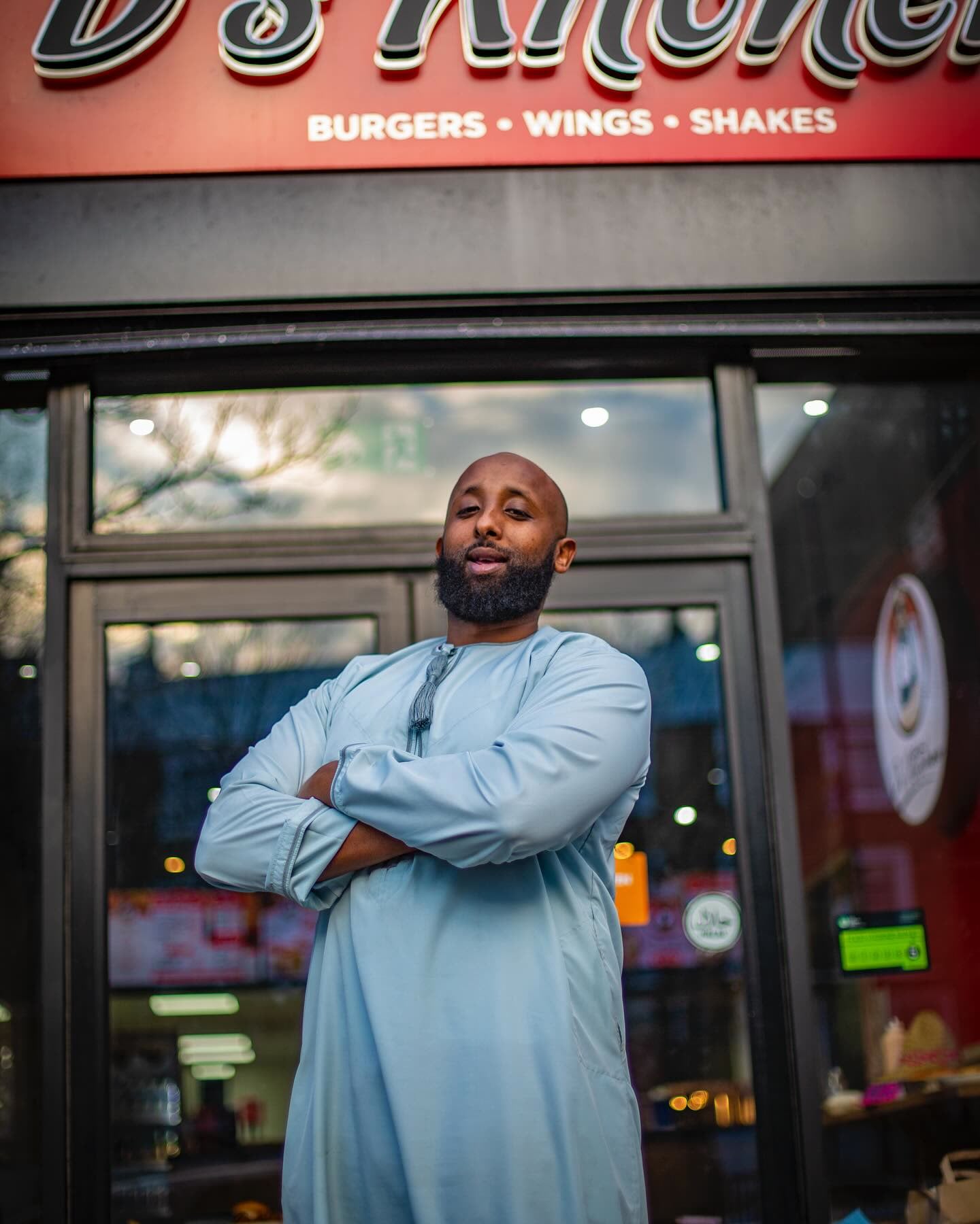 Dirie, arms crossed at the door of a D’s Kitchen shop